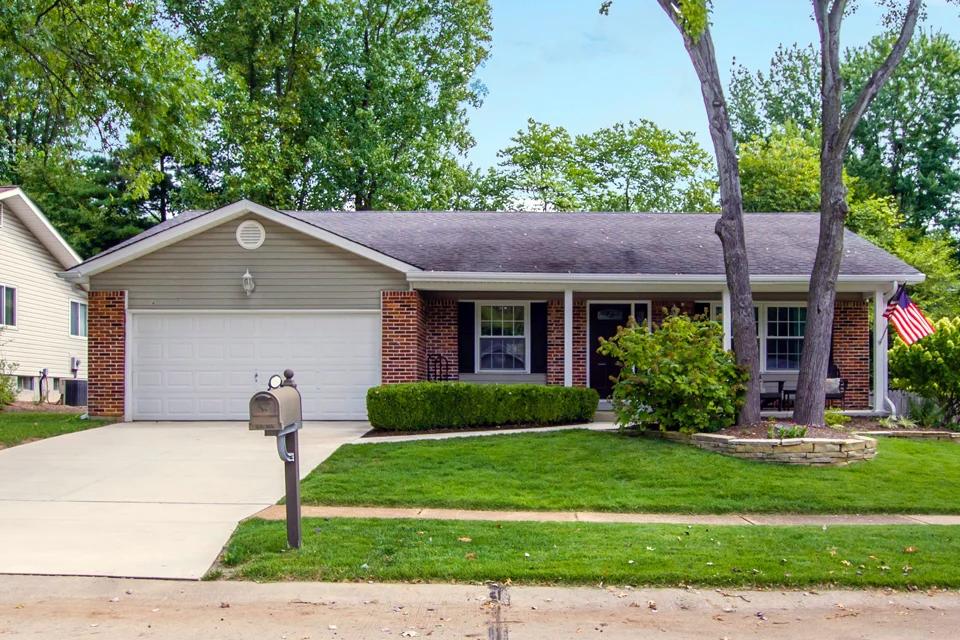 An everyday house viewed from the street which has a bright green lawn, a few bushes and trees in the front yard.
