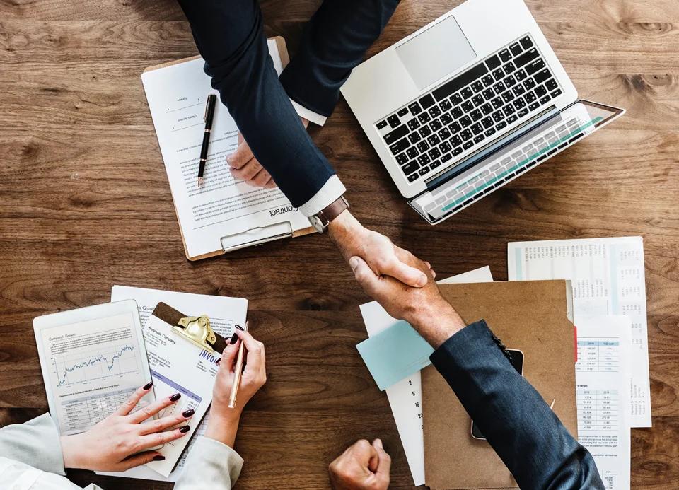 Two men shake hands after what appears to concluding a deal, while a woman watches with a pen in her hand.
